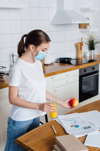 Young woman in medical mask holding glass of orange juice and apple near table with charts and graphs — Stock Photo