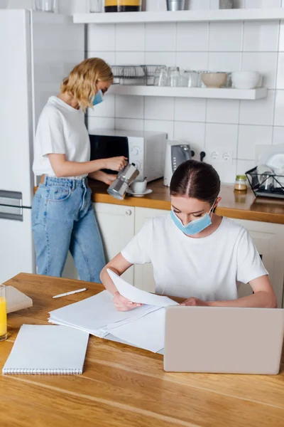 Фрілансер в медичній масці біля ноутбука і сестри на кухні — стокове фото