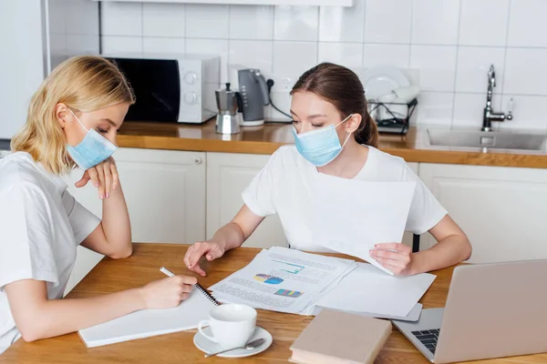 Joven freelancer en escritura de máscaras médicas en cuaderno cerca de hermana, laptop, gráficos y gráficos - foto de stock