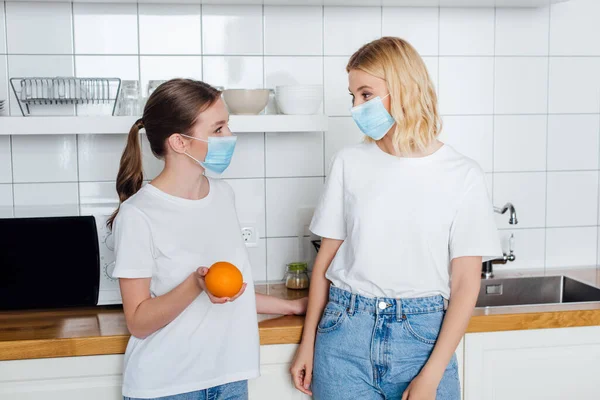 Joven mujer en máscara médica sosteniendo naranja cerca de la hermana - foto de stock