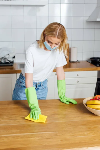 Mulher em máscara médica e luvas de látex segurando pano perto da mesa — Fotografia de Stock