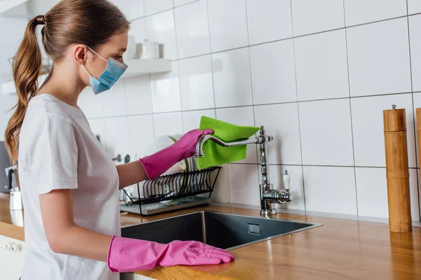 Giovane donna in maschera medica e guanti di gomma che tiene straccio vicino al rubinetto in cucina — Foto stock