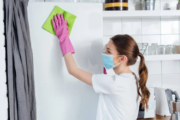 Woman in medical mask and rubber gloves holding rag while cleaning fridge — Stock Photo