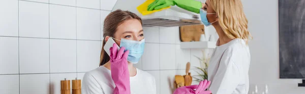 Tiro panorâmico de mulher em máscara médica falando no smartphone perto da cozinha de limpeza da irmã — Fotografia de Stock