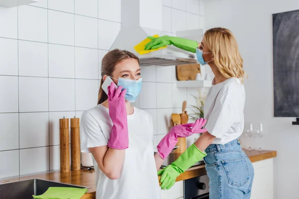 Selective focus of woman in medical mask talking on smartphone near sister cleaning kitchen — Stock Photo