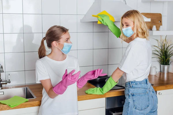 Mulher em máscara médica olhando para irmã em luvas de borracha ao fazer tarefas domésticas — Fotografia de Stock