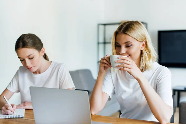 Happy freelancer drinking coffee near sister writing in notebook — Stock Photo