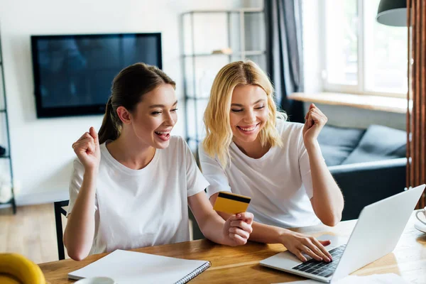 Hermanas alegres compras en línea cerca de la computadora portátil en casa - foto de stock