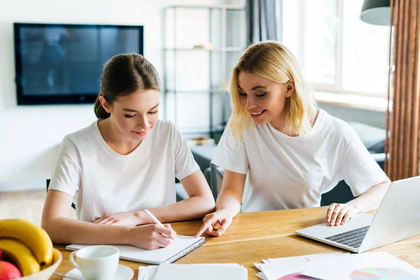 Fröhliche Frau schaut Schwester beim Schreiben in Notizbuch ordentlich Laptop, Online-Studienkonzept — Stockfoto