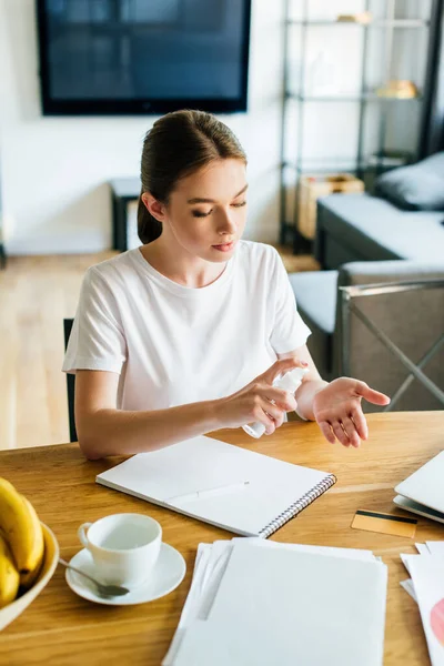 Attractive woman holding bottle with hand sanitizer near notebook and cup on table — Stock Photo