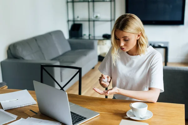 Jeune femme tenant bouteille avec désinfectant pour les mains près d'un ordinateur portable et carte de crédit sur la table — Photo de stock