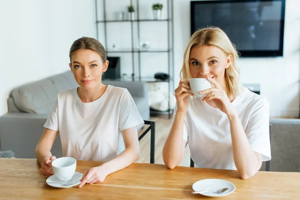 Attraktive Schwestern blicken in die Kamera und halten Tassen Kaffee in der Hand — Stockfoto