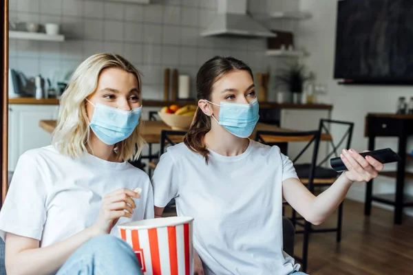 Irmãs em máscaras médicas segurando pipocas enquanto assiste tv em casa — Fotografia de Stock