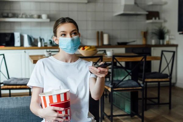 Chica en máscara médica sosteniendo palomitas de maíz mientras ve la película en el sofá - foto de stock