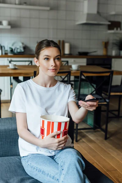 Attraente ragazza che tiene secchio di popcorn e telecomando a casa — Foto stock