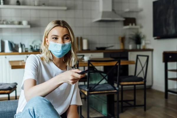 Donna bionda in maschera medica che tiene il telecomando a casa — Foto stock