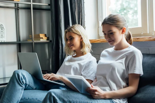 Selective focus of smiling sisters using laptop and digital tablet on sofa — Stock Photo