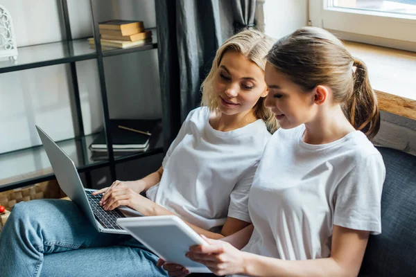 El foco selectivo de la muchacha sonriente que muestra la tableta digital a la hermana con el portátil en el sofá - foto de stock