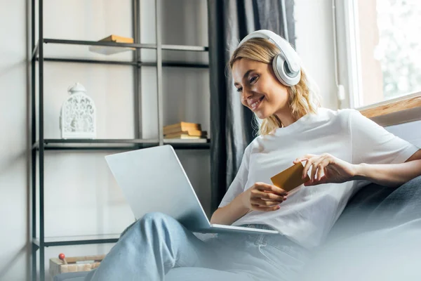 Foco seletivo de mulher sorridente em fones de ouvido usando laptop e segurando cartão de crédito no sofá — Fotografia de Stock