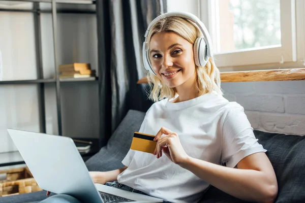 Attraktives Mädchen mit Kopfhörern lächelt in die Kamera, während sie Kreditkarte und Laptop im Wohnzimmer hält — Stockfoto