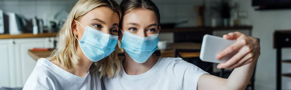 Panoramic shot of sisters in medical masks taking selfie with smartphone at home — Stock Photo
