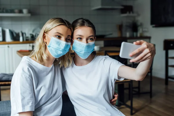 Hermanas en máscaras médicas tomando selfie con smartphone en casa - foto de stock