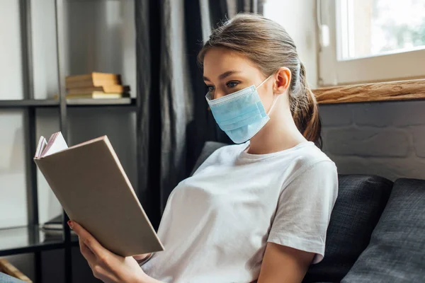 Mujer joven en máscara médica libro de lectura en el sofá - foto de stock