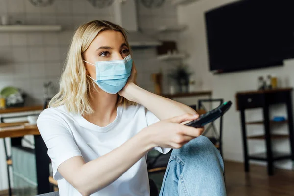 Selective focus of blonde woman in medical mask watching tv at home — Stock Photo