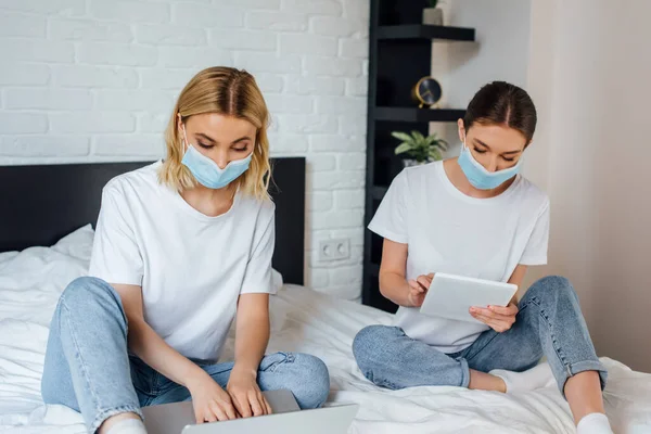 Soeurs dans des masques médicaux à l'aide d'un ordinateur portable et tablette numérique sur le lit — Photo de stock