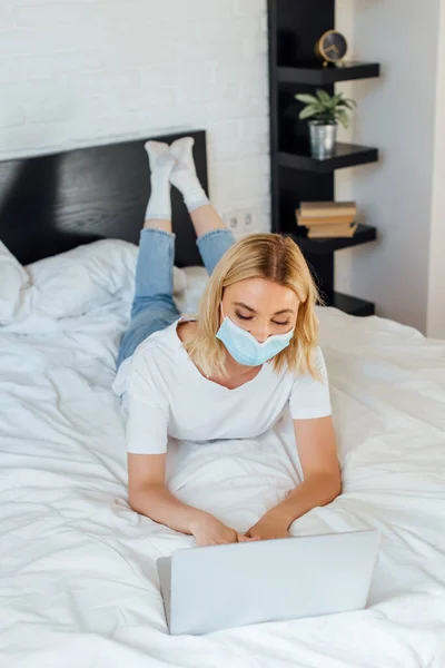 Телеработник в медицинской маске работает на ноутбуке на кровати дома — стоковое фото