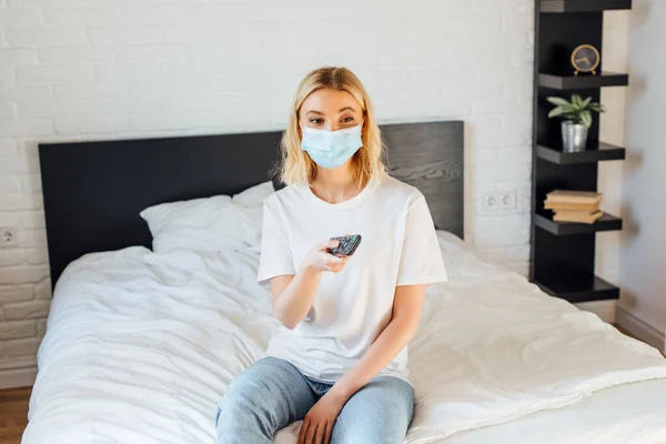 Mujer joven con máscara médica sosteniendo mando a distancia mientras está sentada en la cama - foto de stock