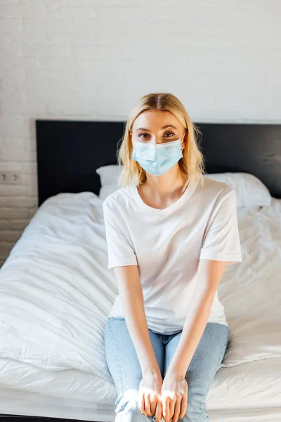 Blonde Frau in medizinischer Maske schaut in die Kamera, während sie auf dem Bett sitzt — Stockfoto
