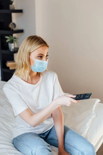 Chica rubia en máscara médica haciendo clic en los canales mientras está sentado en la cama - foto de stock