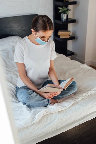 Foco seletivo da jovem mulher em máscara médica livro de leitura na cama — Fotografia de Stock