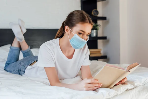 Mujer joven en el libro de lectura máscara médica mientras está acostado en la cama en casa - foto de stock