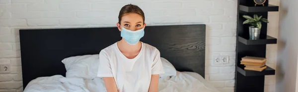 Panoramic crop of young woman in medical mask looking at camera on bed — Stock Photo