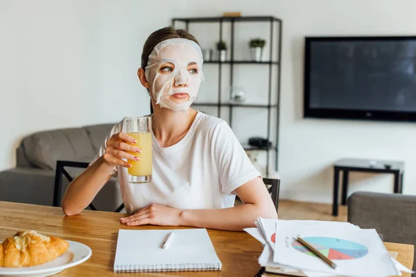 Selective focus of woman in face mask holding glass of orange juice near papers with charts on table — Stock Photo