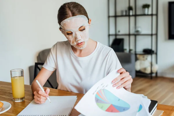 Selective focus of woman in face mask writing on notebook and holding papers with graphs near orange juice on table — Stock Photo