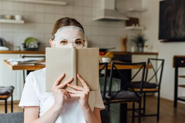 Junge Frau mit Gesichtsmaske blickt in Kamera, während sie Buch zu Hause hält — Stockfoto