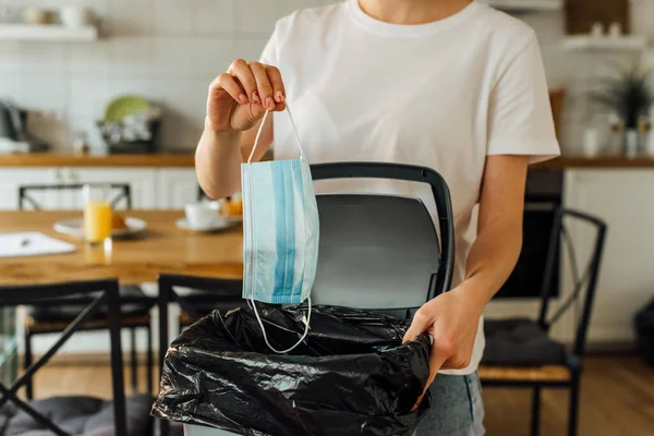 Vista recortada de la mujer sosteniendo máscara médica cerca de bote de basura en casa - foto de stock