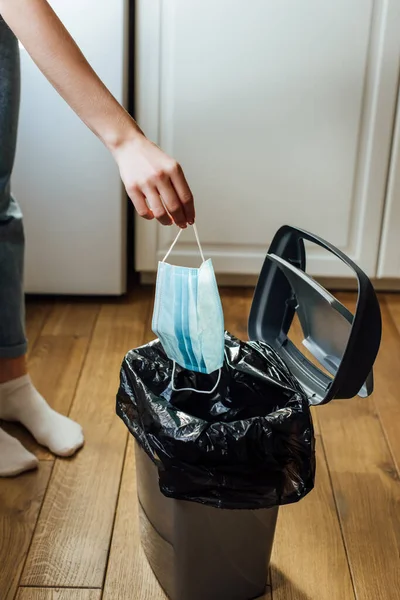 Cropped view of woman holding medical mask near trash can on floor — Stock Photo