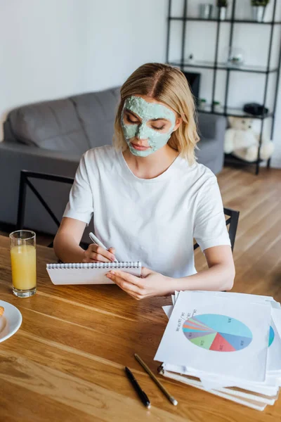 Femme blonde en masque facial écrivant sur un carnet près des papiers et du jus d'orange sur la table — Photo de stock