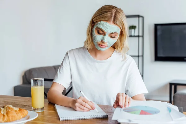 Enfoque selectivo de mujer rubia en mascarilla facial trabajando con documentos y cuaderno cerca de croissant y zumo de naranja en la mesa - foto de stock