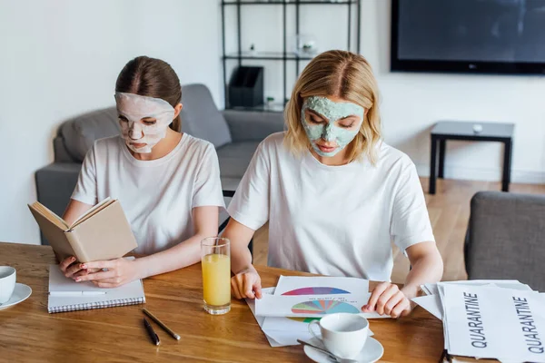 Hermanas en máscaras faciales leyendo libro y trabajando con papeles en la mesa — Stock Photo