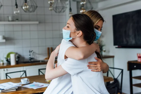 Vista lateral de hermanas con máscaras médicas abrazándose en casa - foto de stock