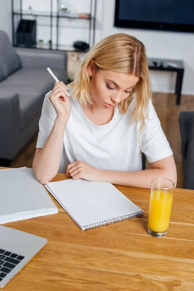 Блондинка держит ручку возле блокнота, ноутбук и стакан апельсинового сока дома — стоковое фото