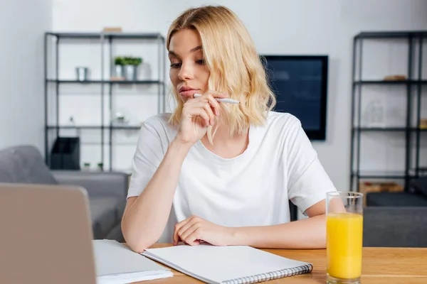 Selective focus of beautiful freelancer holding pen near laptop, papers and orange juice on table at home — Stock Photo