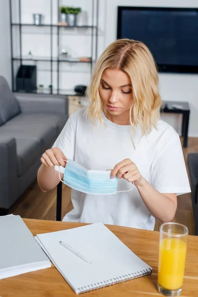 Concentration sélective de la femme blonde tenant un masque médical près du jus d'orange et des papiers sur la table — Photo de stock