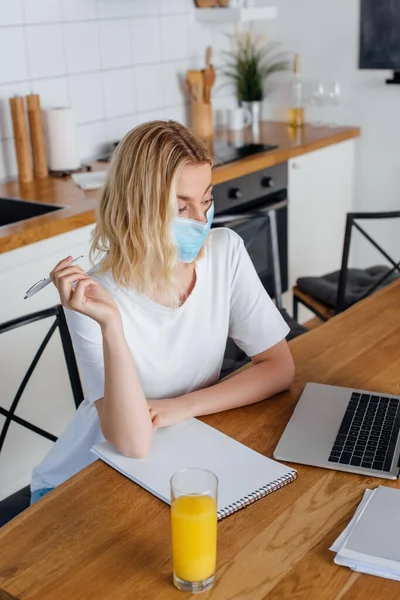 Freelancer em máscara médica segurando caneta perto de suco de laranja, laptop e notebook na mesa — Fotografia de Stock