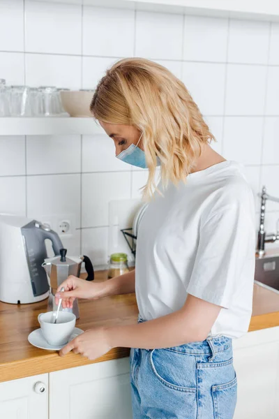 Seitenansicht einer blonden Frau in medizinischer Maske, die in der Küche Kaffee mixt — Stock Photo